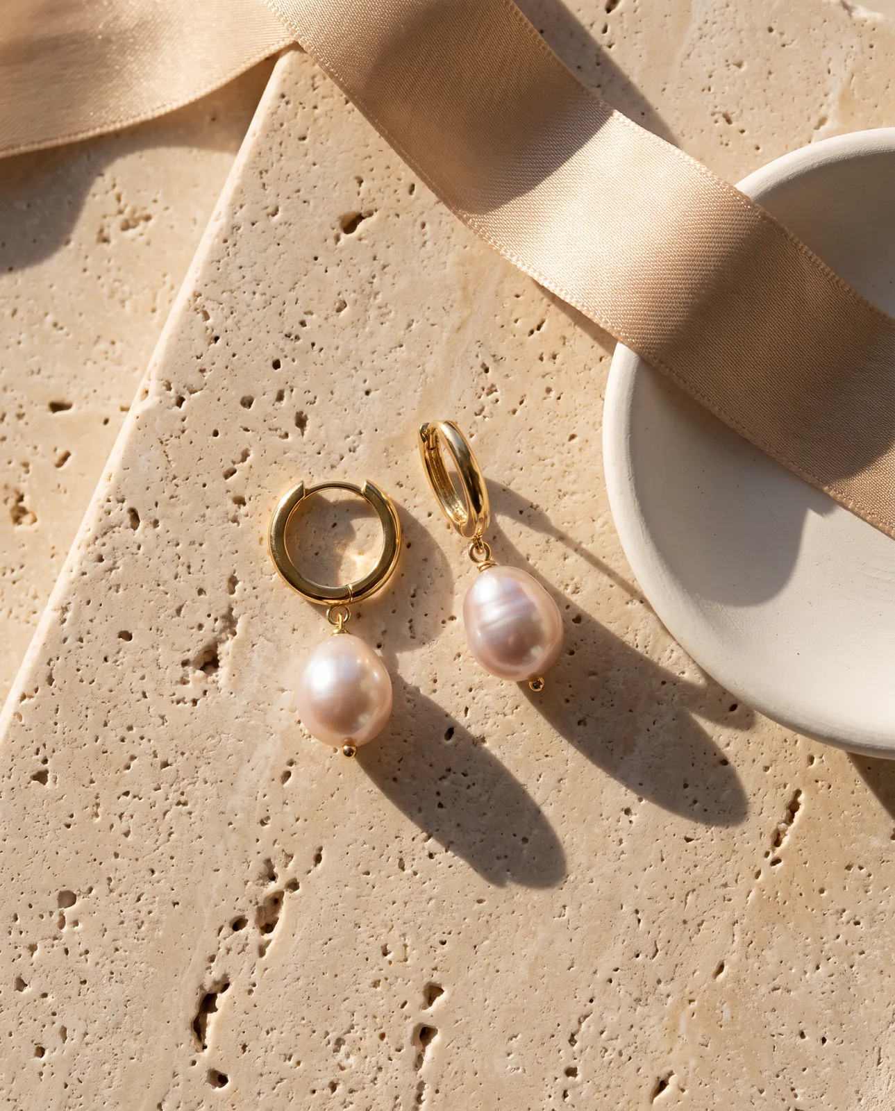 Nano Banana Pro output: gold huggie hoops with pearl drops on travertine with a silk ribbon and a ceramic dish, long shadows
